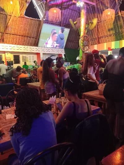 The scene is a lively outdoor restaurant with a thatched roof in Diani Beach, Kenya, located at coordinates -4.276 latitude and 39.592 longitude. The restaurant is dimly lit, with a large television screen playing a sports match in the background. Several patrons are gathered around wooden tables, engaging in conversation. Two men in the background are watching the television screen. A large wooden wheel hangs from the ceiling. The tables are covered with white tablecloths and are decorated with colorful flowers. There is a bar to the left of the scene, where patrons can order drinks and food. The overall atmosphere is relaxed and casual, with a hint of excitement from the sports game on the screen.