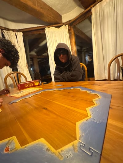 Two people are at a wooden table playing Catan. One person is wearing a dark hoodie and sitting near a window with white curtains. The game board is partially assembled with a blue edge. The Catan box is visible next to the board. Part of the second person's face and hair are visible. The room appears to be lit with warm light.