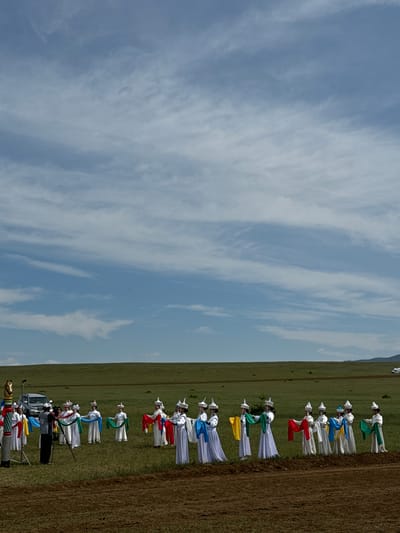 The image depicts an outdoor event in Orongoi, Russia, featuring a group of individuals dressed in traditional white garments with distinctive hats. They are holding colorful sashes in various hues, including blue, green, yellow, and red, and appear to be performing a coordinated dance or ritual in a grassy field. Some of the individuals are holding their sashes in flowing arcs. In the background, a silver SUV is parked. A cameraman with a video camera on a tripod is positioned to the left of the group, actively filming the event. The scene is set against a backdrop of rolling green hills under a cloudy blue sky. A partially tilled field is visible in the foreground.