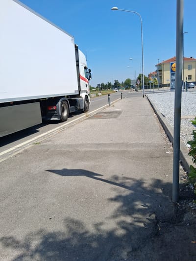 A semi-trailer truck, branded "SCANIA," is parked on the side of a road with its trailer extending towards the left of the frame. The truck is white with a red stripe and the word "SCANIA" is visible on the mud flaps. The truck is on a paved surface, adjacent to a sidewalk. The sidewalk is concrete and runs alongside a gravel parking lot. In the background, there is a LIDL supermarket with its distinctive blue and yellow logo. Several streetlights are visible along the road, which appears to be a main thoroughfare. The sky is clear and blue, indicating a sunny day. The scene is devoid of people. The location is the Via Gandini in Colorno, Italy, in front of the LIDL supermarket.