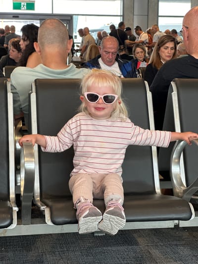 A young child with blonde hair and pink cat-eye sunglasses is sitting in an airport waiting area, holding onto a metal seat divider. The child is wearing a striped long-sleeved shirt and light pink pants, with pink and grey sneakers. Several other people are visible in the background, seated and standing, some facing away from the camera. The environment appears to be an indoor public space with dark grey seats and carpeted floor. An exit sign with a green background and a white figure is visible in the upper left corner.