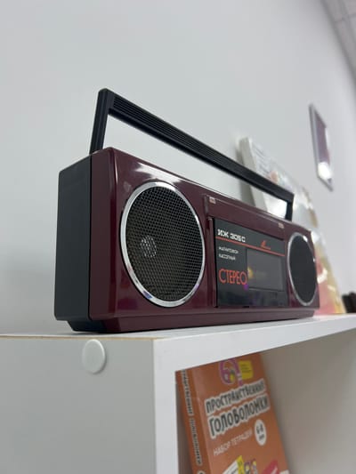A vintage burgundy boombox sits on a white shelf, angled towards the viewer from a low, upward perspective. The boombox features two silver-rimmed speakers, a black cassette deck with "STEREO" printed in red, and a black retractable handle. To the right of the boombox, a blurred book with colorful illustrations and Cyrillic text, titled "Пространственные Головоломки" (Spatial Puzzles), is partially visible on the shelf below. The background consists of a plain white wall and shelf. The scene appears to be indoors, possibly in a home or retail setting. The image is sharp and well-lit, with the boombox being the clear focal point. The overall impression is one of nostalgia and retro technology.
