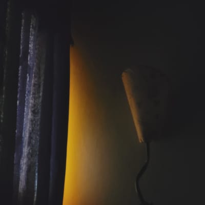 The image depicts a dark indoor scene, likely at night, characterized by a single prominent light source. In the upper left, a partially visible, light-colored lampshade or fixture emits a soft, warm yellow-orange glow. A dark, curved arm extends from this fixture, suggesting it's part of a lamp. This warm light illuminates a horizontal band across the middle of the frame, creating a striking gradient from bright yellow at its base to a softer orange as it fades into the dark background. Below this illuminated area, several dark, horizontal structures are visible, appearing as textured planks, shelves, or possibly blinds. These surfaces show subtle reflections of the yellow light and also exhibit faint blue-ish highlights. The overall atmosphere is quiet and intimate, with no discernible activity or visible text. The image does not contain specific visual cues to indicate its location in San Fernando del Valle de Catamarca, Argentina.