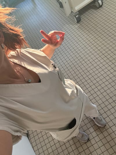 The image depicts a partial, high-angle view of an individual standing indoors, presumably within a medical facility in Tourcoing, France. The subject, likely female, is shown from the chest down, with a portion of reddish-brown hair visible on the left. She is wearing a light beige or off-white V-neck medical scrub top and matching pants, along with grey athletic shoes. A thin necklace with a small, light blue or green pendant is visible around her neck. A dark-colored badge holder or clip is attached to the left side of her scrub top. Her right arm is raised, with the hand forming an "OK" gesture: thumb and index finger creating a circle, while the other three fingers are extended upwards. Strong, directional sunlight enters from the left, casting prominent shadows across the scene. The flooring consists of light grey square tiles with dark grout lines, arranged in a grid pattern. In the upper right background, part of a white mobile medical cart with grey wheels is visible.