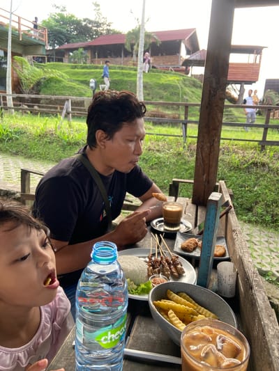 Here's a detailed description of the image:
The image shows a man and a child eating at a table in an outdoor setting. The man is holding a piece of food with a fork, and the child is eating a French fry. The table is covered with various dishes, including satay skewers, French fries, rice, and a glass of iced coffee. There is also a receipt and a bottle of water on the table. The background of the image shows a green, hilly landscape with buildings and other structures. The Nota indicates that this scene is occurring in Tamiajeng, Indonesia.