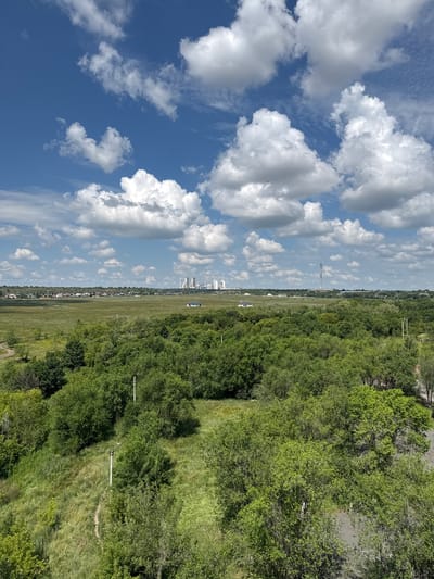 The image depicts a panoramic view of a city landscape under a partly cloudy sky. In the foreground, dense green trees and vegetation dominate the scene, with a pathway winding through the grassy areas. Two white lampposts are visible in the middle ground, adding a touch of urban infrastructure to the natural elements.
Further in the distance, a vast, open field stretches out, dotted with scattered buildings and a single small house with a dark roof. Beyond this, a town with residential buildings and a prominent industrial complex with tall structures and a smokestack is visible on the horizon. The sky is a vibrant blue, adorned with numerous fluffy white clouds, some casting shadows on the landscape below. The overall impression is one of a typical day in Novotroitsk, Russia, a city known for its industrial activity, blending urban development with natural open spaces.