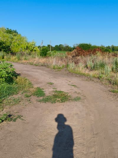 A shadow of a person wearing a hat is cast on a dirt road in Psilske, Ukraine. The road is flanked by dry grass, green vegetation, and trees. The sky is clear and blue.
