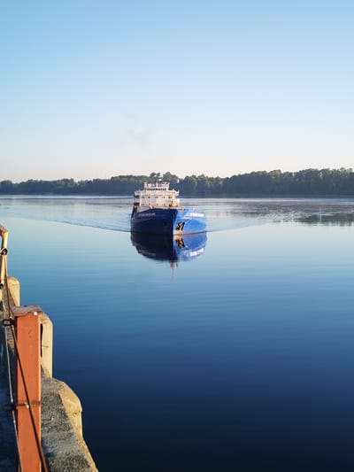 The image depicts a serene, wide river with calm, blue waters reflecting the clear sky. A blue cargo ship, identified by the lettering "ЕУГЕНИИ ГРАДОВСКИИ" on its side, is navigating down the river, leaving a gentle wake behind it. The ship has a white superstructure with several decks and windows. In the background, a dense line of green trees forms the riverbank, with a faint haze or mist rising from the water near the trees. On the left side of the foreground, a portion of a concrete structure, likely a pier or dock, is visible with rusted metal railings. The lighting suggests it is early morning or late afternoon, with the sun casting a soft light on the scene. The location is identified as the city of Bol'shie Mkhi, Russia, a place known for its riverine landscape.
