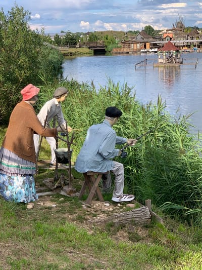 In the city of Malakhovetski Sel'ski Sovet, Belarus, an outdoor scene depicts three mannequins engaged in fishing by a lake. The mannequin on the left is dressed in a brown knitted cardigan, a long blue and white floral skirt, and a pink knitted hat, and is standing with its hands resting on a wooden support. Next to it, a mannequin in a light-colored long-sleeved shirt and cap is holding a fishing rod. The third mannequin, seated on a wooden stool and wearing a blue collared shirt, gray trousers, and a black cap, is actively fishing with a rod and reel. The mannequins are positioned amidst tall green reeds and grass. The lake extends into the background, where a picturesque village with wooden houses and a bridge is visible across the water. The sky is partly cloudy, with fluffy white clouds against a pale blue backdrop. The overall atmosphere suggests a tranquil, rural setting.