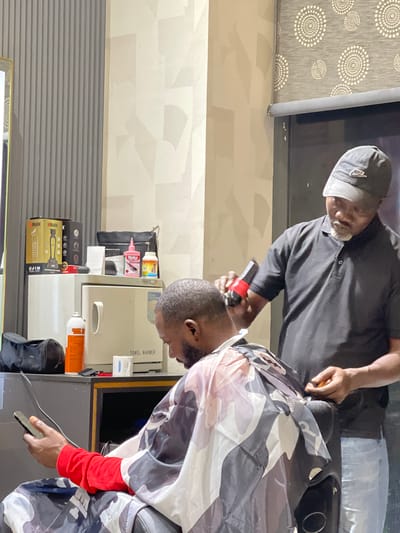 In a barbershop located in Guzape, Nigeria, a barber is seen trimming the hair of a client. The barber, wearing a black polo shirt and a grey Nike baseball cap, is focused on the task, holding a cordless hair clipper in his right hand. The client, seated and draped in a camouflage cape, is looking down at his mobile phone, which he holds in his left hand. The background of the barbershop reveals a grey, vertically slatted wall on the left, a beige wall with a geometric pattern, and a window covered by a roller blind with a circular pattern. On a shelf in the background, several items are visible, including two boxes of "WMARK" hair clippers, a pink bottle of "Medi+Plus," a small orange bottle, a white paper towel, and a black bag. A white towel warmer with the label "TOWEL WARMER" is also present on the shelf, and a can of "NOW" spray is on the counter below. The overall scene captures a moment of personal grooming within a typical urban barbershop setting.