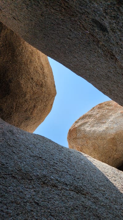 The image captures a ground-level view looking upwards through a narrow opening formed by large, textured rock formations. The rocks appear to be natural, with a rough, granular surface. A patch of clear, blue sky is visible through the opening. The color of the rocks varies from a darker, gray-brown to a lighter, tan shade. The scene evokes a sense of being enclosed within a rocky landscape. The location is Phuoc Dinh, Vietnam.