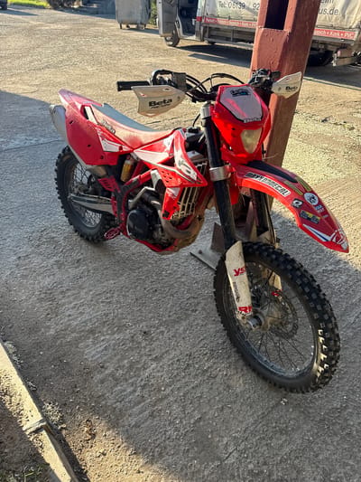 The image captures a red Beta motorcycle parked near a weathered, rectangular wooden post, likely a support structure, set against a backdrop of textured concrete. The motorcycle shows signs of use, evidenced by the dirt and wear on its components. The surroundings include a vehicle partially visible in the background, and a glimpse of what appears to be an urban or semi-urban environment. The presence of a wooden post and concrete suggests an outdoor setting with a mix of structural elements and natural light. It cannot be determined the location is in the city of Bratya Daskalovi, Bulgaria from the image provided.
