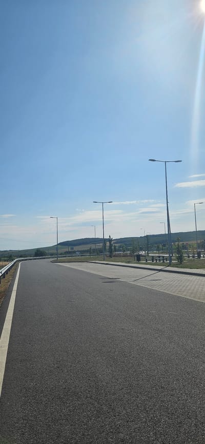 The image depicts a wide, asphalt road curving to the left, with a white line marking the edge of the travel lane on the left side. The road surface is textured and dark grey. Beyond the road, on the right, is a paved area with a light grey, interlocking paver pattern. Several tall, modern streetlights with horizontal fixtures are visible along this paved area. In the background, beyond the paved area, are scattered trees and picnic tables with benches, suggesting a rest stop or park area. Further in the distance, rolling hills covered in greenery and trees form the horizon under a clear, bright blue sky. The sun is visible in the upper right corner, casting a bright glare and lens flare. The scene appears to be in a rural or semi-rural setting, likely on the outskirts of Hidasnémeti, Hungary, given the context provided. No people are visible in the image.