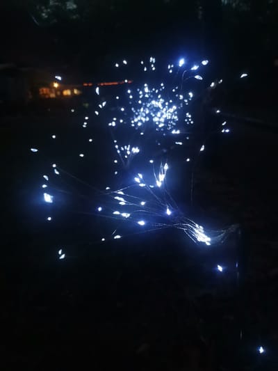 The image depicts a close-up, low-light view of a string of small, white LED lights that appear to be arranged in a dandelion-like or firework-like cluster. The lights are attached to thin, dark wires that spread out from a central point. Many of the lights are bright and emit a cool white glow, with some appearing slightly blurred due to motion or focus. In the background, out of focus, are hints of a building with illuminated windows, suggesting an urban or suburban setting at night. The overall impression is one of decorative outdoor lighting. The location is the city of Asprovalta, Greece.