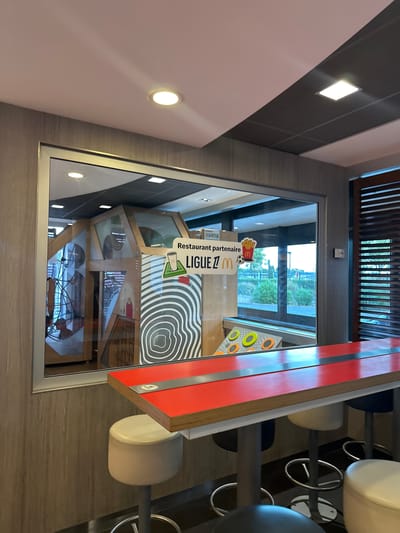 The image shows the interior of a McDonald's restaurant, likely located in Narbonne, France, based on the "Ligue Z M" signage which is a common feature in French McDonald's restaurants that partner with youth sports leagues. The main focus is a glass-enclosed play area for children, visible through a large window. Inside the play area, there is a wooden structure with various climbing elements and decorative panels. Outside the play area, in the foreground, a bright red table with metallic accents is positioned with several bar stools of various colors (white, black, and possibly beige). The table has circular indentations, suggesting it might be interactive. The walls of the restaurant are paneled with a textured material that resembles wood grain. The ceiling features modern lighting, including recessed circular and rectangular fixtures. Through the window of the play area, a glimpse of the outdoors can be seen, with green foliage and a fence suggesting an exterior area, possibly a parking lot or garden. No people are clearly visible in the immediate foreground, but reflections in the glass of the play area suggest the presence of individuals inside. The overall scene depicts a typical family-friendly dining and play environment within a fast-food establishment.