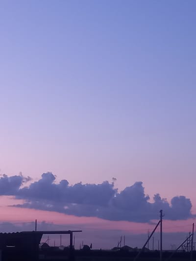 The image captures a twilight sky over an industrial or semi-rural area in Қозыкеткен ауылдық округі, Kazakhstan. The sky transitions from a pale lavender at the top to a soft pinkish hue near the horizon. A line of dark, irregularly shaped clouds stretches across the mid-ground. Below the clouds, silhouetted against the sky, are various structures. On the left, a dark, angular roofline of a building is visible. Towards the center and right, a series of utility poles, some with diagonal supports, are prominent, suggesting electrical infrastructure. The overall scene is characterized by muted colors and stark silhouettes, indicative of the fading light. No people are visible in the frame.