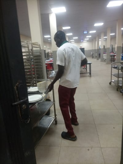 In a spacious hall with tiled floors and numerous pillars, a man stands near a utility cart piled with plates. He is dressed in maroon pants, a white shirt, and casual footwear. The scene suggests a dining or catering area, indicated by the arrangement of tables, chairs, and additional utility carts further down the hall. Bright ceiling lights illuminate the space. Part of a dark door is visible in the foreground.