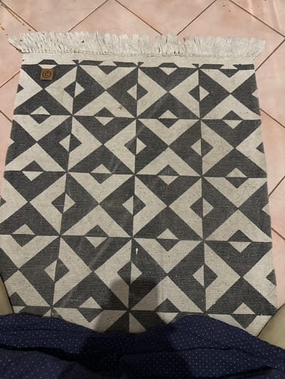 An overhead, full-frame shot shows a dark grey and off-white geometric patterned rug with a fringed edge. The rug is positioned on a tiled floor. In the upper left corner of the rug is a circular brown tag with a stylized "C" logo. Some light-colored fibers are scattered on the rug and the floor. The bottom of the image shows a dark blue fabric with a white polka dot pattern. The scene does not contain any identifying features or context to place it in Kalitino, Russia, or any specific city.