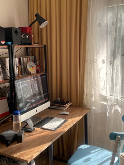 The image shows a desk setup in what appears to be a home office or study area. The desk is made of wood and is positioned against a wall with light brown blinds and sheer white curtains with a floral pattern. Sunlight streams through the curtains, suggesting it is daytime.
On the desk, there is an iMac computer with its screen displaying what looks like a software interface with various icons and windows. To the left of the computer, there is an Apple keyboard and a mouse. A small black desk lamp with a flexible neck is positioned on a shelf below the desk. Next to the lamp are two red speakers and a black subwoofer.
A shelf above the desk holds a stack of books, some of which have Turkish titles like "akilgözü," "NOROPSIKAP," "FITRATA UYGUN ÇOCUK," "ZİNDE REHBER," "HAFIZA KOÇU," and "TEKNOPOLIS." There is also a colorful circular object, possibly a fan or a decorative item, and a small golden figurine. Further to the right, on the desk, is a clear plastic bottle with a blue cap and some contents inside, alongside a black electronic device.
The overall impression is that of a functional workspace, possibly for creative or technical work, with personal items and books indicating the user's interests. The presence of Turkish text on the books strongly suggests the location is in Turkey, and the context provided mentions Başakşehir, Turkey.