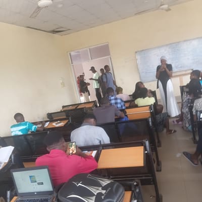 This image depicts a group of people in a classroom setting in Wukari, Nigeria. The room is equipped with desks and chairs, suggesting an educational environment. Several individuals are seated at desks, some looking towards a whiteboard at the front of the room, which appears to have writing on it, though it's mostly illegible. Others are interacting with electronic devices, such as laptops and smartphones. One person is holding up a smartphone, displaying what appears to be an image of people.
The individuals are dressed in casual attire, with some wearing head coverings. The lighting suggests it is daytime, likely indoors, as there are overhead lights visible and the natural light appears diffused. The atmosphere seems to be one of a lesson or presentation in progress, with a mix of focused attention and casual engagement. The overall mood is neutral, conveying a typical day in an academic institution. No specific weather conditions are discernible, but the indoor setting implies protection from external elements.