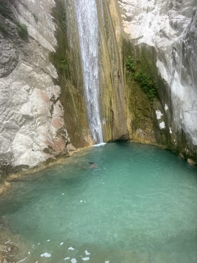 A person is partially submerged in the clear, turquoise water of a pool at the base of a waterfall. The waterfall cascades down a tall, rough rock face that is partially covered in green moss and streaks of brown and white. The pool is surrounded by rocky terrain, with sheer rock walls forming a natural enclosure. The water appears calm and shallow near the edges, with sunlight dappling the rocky bottom visible in some areas. The scene suggests a natural, outdoor setting, likely a secluded grotto or a less-trafficked waterfall location.
