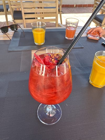 This image captures an outdoor dining scene, likely a café or restaurant patio, under bright daylight. The central focus is a large, stemmed glass filled with a shimmering, deep red beverage, which appears to be a cocktail or a fruit-based drink. The drink is garnished with several pieces of dried red fruit, possibly strawberries or raspberries, and a colorful mixture of dried floral petals, including blue, pink, and yellow tones. A black drinking straw is positioned within the glass.
The glass is situated on a dark grey, rectangular placemat with a noticeable woven texture, which rests on a light-colored wooden table. To the upper left of the main drink, a pair of dark tortoiseshell-patterned sunglasses with dark lenses is placed on the table, partially overlapping another dark grey placemat.
Further on the table, towards the upper right, a smaller clear glass contains a bright orange liquid, consistent with orange juice. Adjacent to it, another clear glass holds a lighter, reddish-orange liquid. A person's hand, displaying a light skin tone, is visible on the far right, holding an orange mobile phone. The phone's screen shows a circular graphic. A black object, possibly a pen, is partially visible near the phone. In the lower right foreground, a third clear glass containing a bright yellow-orange liquid is partially visible.
The background reveals a patio setting with multiple slatted wooden chairs featuring dark seat cushions, arranged around similar tables. Beyond the immediate seating area, a light-colored building wall with horizontal reddish-brown bands (possibly tiles or bricks) is visible, topped by a white coping or railing. The ground surface appears to be light-colored concrete or paving.
The image does not contain any discernible visual cues or identifying markers to confirm the specific location as Basel, Switzerland.