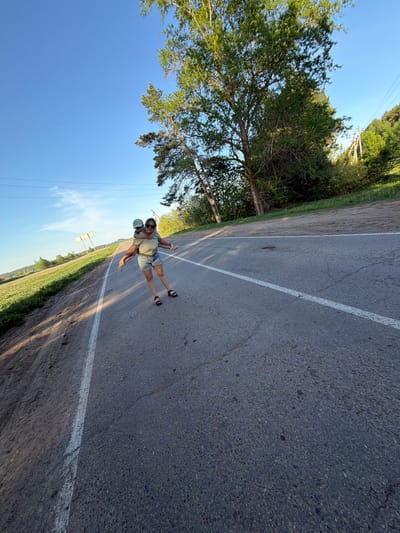 A woman is giving a piggyback ride to a child on an asphalt road with white lane markings. The woman is wearing sunglasses, a light-colored striped shirt, and denim shorts. The child is wearing a blue cap, a light-colored shirt, and denim shorts. The road is surrounded by trees and fields. The sky is blue. The photo was taken in Foki, Russia.