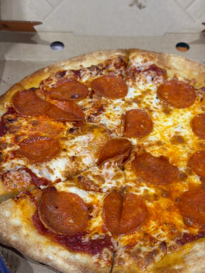 A close-up, overhead shot shows a pepperoni pizza in a cardboard box. The pizza is cut into slices, and the crust is golden brown and slightly puffy. The toppings include melted cheese and round slices of pepperoni. The pizza appears to be freshly made and ready to eat. The box is open, revealing the pizza within. The lighting is bright and even, highlighting the textures and colors of the pizza. There are no people or other objects visible in the frame. The background consists of the inside of the pizza box.