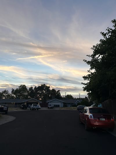 The image depicts a suburban street scene at dusk in Citrus Heights, California. The sky is filled with wispy, colorful clouds, suggesting a sunset or sunrise with dramatic lighting. Tall trees with dark green foliage line the street and partially obscure the houses, which appear to be single-family homes with pitched roofs.

Several vehicles are parked along the asphalt road, including a red Nissan Sentra, a white SUV, and a black sedan. The Sentra has a license plate visible that reads "7VNN874". In the foreground, a black trash bin is visible. The overall impression is one of a quiet evening in a residential neighborhood. The lighting and the scattered clouds create a peaceful and serene atmosphere. There are no people visible in the image. The time of day is clearly indicated by the soft, warm light illuminating the clouds and casting long shadows.