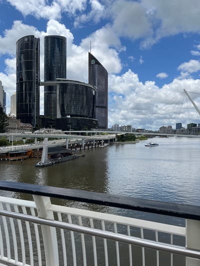 This daytime image presents a panoramic view of a modern cityscape alongside a wide river, likely the Brisbane River in South Brisbane, Australia. The scene is captured from an elevated perspective, possibly from a ferry or a riverside structure, indicated by the white and metallic railing visible in the foreground on the right.

The sky is a bright blue, adorned with fluffy white cumulus clouds, suggesting clear and sunny weather. The wide river, exhibiting gentle ripples, dominates the midground and stretches into the distance. A white and dark passenger boat or ferry can be seen moving down the river in the background, while a smaller, dark boat is docked closer to the riverbank.

Along the left bank of the river, an active urban waterfront features an elevated white track or walkway, possibly a monorail or pedestrian bridge, running parallel to the water. Beneath this, a series of dark piers or docks with multiple posts extend into the river.

The left side of the image showcases a distinctive collection of contemporary skyscrapers. Most prominent on the far left is a dark, curved glass building with the text "THE STAR" clearly visible on its facade. Adjacent to it are other dark, cylindrical high-rises. Further back and slightly to the right, a taller, dark skyscraper with an angular, pointed top stands out against the blue sky. In the upper background, a white cable-stayed bridge, characterized by its prominent cables, connects to more distant urban development.

The image captures the dynamic essence of South Brisbane, highlighting its modern architecture and the vital role of the river in its urban landscape. The primary subjects are the city's infrastructure, its river, and the contemporary buildings. No specific event or human activity is central, rather it depicts the general ambiance of a vibrant city during the day.