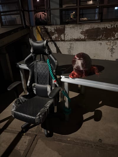 A gaming chair, camouflaged with grey and black patterns, is positioned on a concrete floor in front of a dark, worn table. A thick, green rope is tied to the chair's base and extends upwards, partially obscuring the chair's back. On the table, a red, translucent object resembling a head or mask is placed to the right of a dark, rectangular surface. In the background, a grimy, industrial-looking wall with peeling paint serves as a backdrop. Above the wall, through a window, a blurred image of hands is visible. The scene appears to be in an outdoor, possibly abandoned, urban setting.