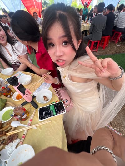 An overhead selfie captures a young woman at an outdoor gathering in Tay Son Commune, Vietnam. She is positioned at the top of the frame, lying on her side or leaning over a table, wearing a light-colored, possibly off-white, formal dress with sheer fabric. She makes a peace sign with her right hand and looks playfully towards the camera.
The scene below her shows a lively outdoor event with multiple people seated at tables covered with yellow tablecloths, likely enjoying a meal. Many attendees are dressed in suits or semi-formal attire, while others wear more casual clothes, including red and white traditional-looking garments. Red, white, and blue fabric decorations are visible in the background, suggesting a festive occasion. The ground appears to be covered in artificial grass.
On the table in front of the main subject, various items are scattered: empty and partially eaten bowls, chopsticks, a pink portable camera or mini tripod, glasses, and saucers with dips. A white smartphone is prominently displayed, showing an app interface. Visible text on the phone screen includes "Ruby," "MAPPER," "Your mapper, rank is #1801," "13.5K," "$0," "$29.84 $10.89," "Get 29 PATTN," and "Withdraw ATTIK." The bright lighting suggests it is daytime. The overall atmosphere appears social and celebratory.