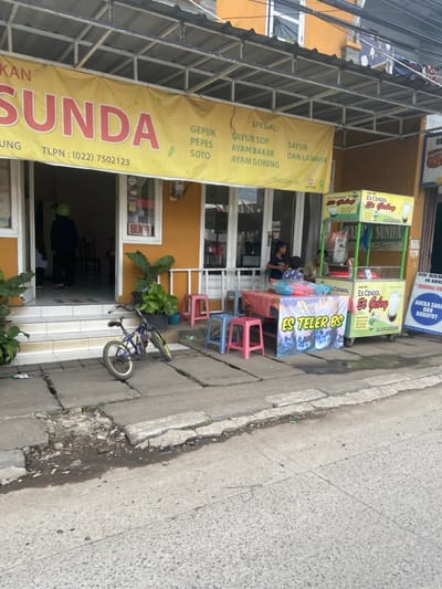 The image depicts a street scene in Bojongsoang, Indonesia, located at latitude -6.979 and longitude 107.634. In the foreground, there is a small, blue bicycle parked on the sidewalk. To the right of the bicycle, there is a small stand with a green frame. The stand is selling "Es Cendol Si Geloy" and "Es Teler Bs." There is also a sign that reads "Aneka Snack dan Buruyot." The stand has an opaque plastic container filled with a beverage and a clear plastic container with a red lid, which may contain a different beverage. Two people are sitting at a table with a red tablecloth. They are likely waiting for their orders.
In the background, there is a building with a large, yellow sign that reads "Sunda" in red letters. The sign lists various menu items in Indonesian, including gepuk, pepes, soto, sayur sop, ayam bakar, and ayam goreng. There is a person in the doorway of the building. It is not possible to see the gender of the person or what they are doing. The sidewalk is made of concrete blocks. The street is paved with asphalt. There are several green plants in pots in front of the building. The building is a yellow color and has a white awning. There is a white roof above the awning. The scene is a typical street scene in an urban area of Indonesia, with a mix of businesses and residential dwellings.