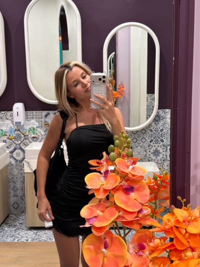 This media file shows a young woman taking a selfie in a bathroom. She is wearing a black dress and holding a silver smartphone. The bathroom has ornate blue and white patterned tiles on the walls and a wooden floor. There are two oval mirrors with white frames on the wall. In the foreground, there is a bouquet of orange orchids. The lighting suggests it is daytime. The woman appears to be in good spirits. The image is taken from a low angle, looking up at the woman.