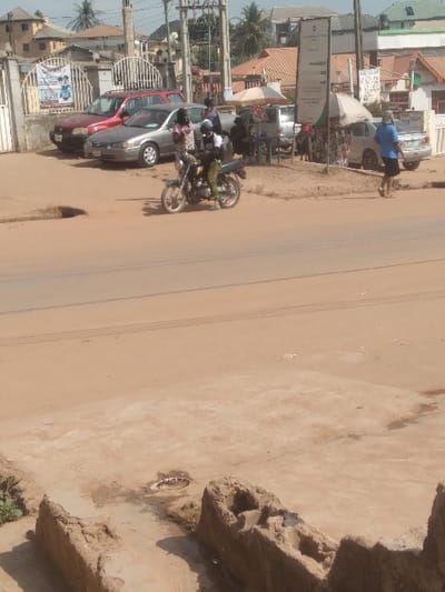 The image depicts a street scene in Agege, Nigeria. In the immediate foreground, an uneven, sandy-brown ground contains two irregular, dilapidated concrete or mud structures and a circular metal drain cover. The midground features a wide, unpaved reddish-brown dirt road with discernible tire tracks and a distinct pothole near the left curb. A person on a motorcycle, wearing a dark top, light-colored camouflage pants, and a dark helmet, is seen riding leftward on the road. Adjacent to the road, near a white metal gate, a red sedan and a silver sedan are parked. Three individuals stand near these vehicles: two females in patterned dresses and one male in a light-colored shirt. Further along the roadside on the right, several individuals are positioned under two light-colored market umbrellas, indicative of a vendor setup, with merchandise visible. Another silver sedan is parked near this area. A tall, white signpost and multiple utility poles with complex wiring intersect the upper portion of the scene. Various residential or commercial buildings with roofs of red, green, and brown hues are present in the background. On the far right, a female in a light blue top walks leftward, away from the camera. The scene is illuminated by what appears to be bright, diffused daylight.