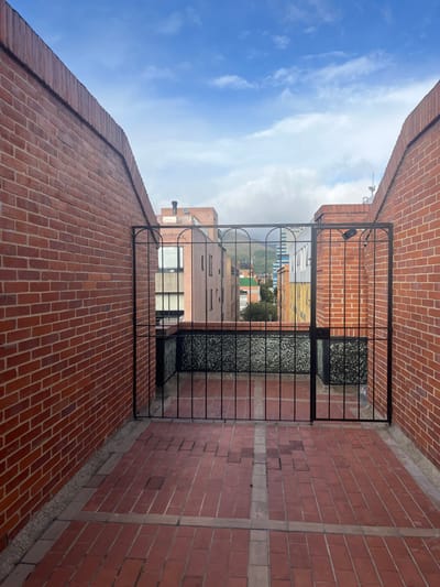 This image depicts an elevated outdoor terrace or rooftop in Bogota, Capital District, Colombia. The foreground is a patterned floor paved with rectangular red bricks, some exhibiting minor surface irregularities and alignment inconsistencies. The space is enclosed on the left and right by two parallel, vertical walls constructed from red bricks, each approximately 3 meters high, featuring a slightly sloped uppermost course. Centered between these walls is a black, wrought-iron gate, approximately 2 meters high, composed of vertical bars and decorative semicircular arch details at its top, appearing to be in a closed position.

Beyond the gate, a low parapet wall with a dark, mottled, possibly aggregate or textured surface is visible. The background reveals an urban landscape: a dense arrangement of multi-story residential and commercial buildings of varying heights and architectural styles, predominantly in shades of beige, brown, and white brick and concrete. A dark industrial chimney is visible on one mid-ground structure. In the distant background, a prominent mountain range, partially obscured by white cloud formations, defines the natural horizon. The sky overhead is a mix of clear blue and scattered white stratocumulus clouds. No individuals, overt actions, or specific interactions are observed within the scene.