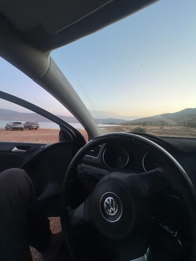 A view from inside a car, likely a Volkswagen, with the steering wheel prominently visible. The open car door offers a landscape view of what appears to be the Bin El Ouidane region in Morocco. Two vehicles, a Toyota truck and a silver sedan, are parked on a dirt area near a body of water. The distant landscape includes arid hills. The sky is light, suggesting either dawn or dusk.