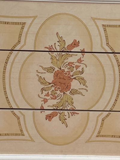 The image shows a close-up of a decorative panel, likely part of a piece of furniture or a wall. The panel features an ornate floral design painted in shades of reddish-brown and muted green, set against a pale cream background. The central element is a large, detailed flower resembling a rose, with layers of petals. Flanking this central bloom are smaller floral elements and stylized leaves. The entire design is framed by an oval border in a soft yellow hue, which is further enclosed by decorative framing elements with a striped pattern. The panel appears to be divided vertically by thin, dark lines, suggesting it might be a cabinet door or a section of a larger patterned surface. The lighting is even, indicating an indoor setting, and there are no visible shadows to suggest a specific time of day or weather conditions. The overall aesthetic is traditional and decorative, with no people or action depicted. There is no visible text in the image.