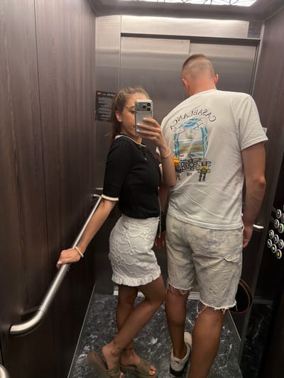 In an elevator with dark wood-paneled walls and a stainless-steel ceiling, a young Caucasian woman with dark hair pulled back in a ponytail and a man with a buzzed haircut are taking a photo with a smartphone. The woman is wearing a black short-sleeved top with a white collar and cuffs, and a white lace skirt. She has a pearl bracelet on her left wrist and is holding the smartphone in her right hand. Her left hand is resting on a silver handrail. The man is wearing a white t-shirt with a graphic print on the back and light-colored denim shorts with frayed hems. He is standing to the right of the woman, with his back to the camera. The elevator also has a panel with illuminated buttons and a dark circular object on the floor to the right. The floor of the elevator is patterned with dark gray and black. The overall scene appears to be a candid moment captured in a private or public building. The location of Hisarya, Bulgaria, is not visually depicted in the image and cannot be confirmed.