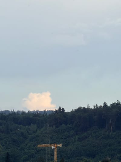 The image captures a cloudy sky above a densely forested hillside. In the foreground, a construction crane with yellow and gray components is visible, partially obscured by the trees. A tall, lattice tower, possibly for electricity transmission, stands near the crane. The environment appears to be a natural landscape with a human-made construction element. The image is taken from a distance, showing the scale of the natural terrain relative to the industrial equipment.