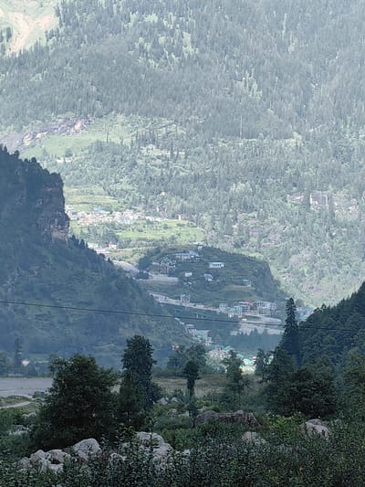 The image shows a mountainous landscape in Palchan, India, characterized by steep, tree-covered slopes. In the midground, a cluster of buildings, possibly a village or resort, is visible nestled on a hillside. A winding road is also discernible, leading through the terrain. The foreground features rocks and dense green foliage. The overall impression is a remote, scenic environment with signs of human settlement integrated into the natural landscape.