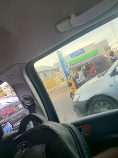 A street-level view from inside a vehicle looking out towards a traffic scene in Karu, Nigeria. The sky is overcast. In the foreground, the interior of the car is visible, including a black backpack on the seat and a seatbelt. Through the side window, a white car is in the right lane, partially obscuring a yellow auto-rickshaw (Keke Napep) in the left lane. The auto-rickshaw has a brown roof and is carrying at least three passengers, including a driver wearing a white shirt and a woman in a red top. A man wearing glasses and a striped shirt stands to the right of the auto-rickshaw. Behind the auto-rickshaw is a building with a green stripe and a blue "Fidelity" logo. Further in the background, a yellow fence encloses a property with a building featuring multiple windows. Another dark-colored car is visible in the far left lane. The overall impression is of a typical urban traffic situation.