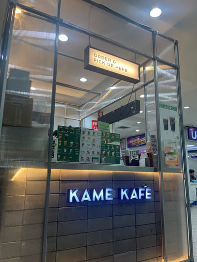 The image shows the exterior of a coffee shop called "KAME KAFE" in Nong Pa Khrang, Thailand. The shop is enclosed by a structure made of metal framing and translucent corrugated panels. Inside, a menu board displays various coffee and tea options, including iced and hot coffee, Thai tea, matcha, and chocolate latte. Prices are visible, such as "55.-" and "70.-". A sign above the counter reads "ORDER & PICK UP HERE".
The lighting suggests it is daytime, with warm light coming from inside the shop and the ceiling lights. The overall aesthetic is modern and minimalist, with a dark tiled wall behind the "KAME KAFE" sign which is illuminated with blue neon lights. The scene appears to be set in a public area, possibly a mall or market, given the surrounding environment and the presence of other signage in the background. One person is partially visible behind the counter, appearing to be a staff member. The atmosphere seems calm and inviting, typical of a casual coffee shop setting.