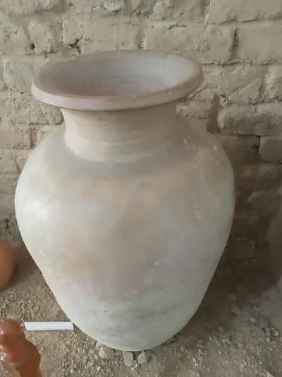 A large, unglazed clay amphora sits on a dusty floor in front of a rough brick wall. The amphora is light brown and has a rounded body that tapers to a narrow neck and a wide, flared opening. The surface of the amphora is uneven and shows some smudges and dust. The brick wall behind it is made of rough, rectangular bricks that are unevenly laid and appear weathered. The floor is covered with dirt and small pebbles. In the lower left corner of the image, part of a small, reddish-brown clay object is visible, possibly a fragment or a small statue. A white label with text is also visible on the floor in front of the amphora. The overall scene suggests an archaeological or historical exhibit. This scene is located in the city of Angren, Uzbekistan.