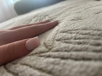 The image shows a close-up, low-angle shot of a person's hand with light pink, square-shaped nails resting on a textured, light-colored surface, possibly a blanket or carpet. The background is blurred, showing hints of a bright light source, possibly a window with sheer curtains, and a darker, out-of-focus object that could be furniture. The scene appears to be indoors. The location in Slobozia-Dușca, Moldova, is not ascertainable from the visual information provided in the image.