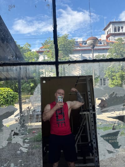 A man is captured in a selfie taken in a large, grimy mirror or window. He is seen from the chest up, flexing his right bicep while holding a smartphone to take the photo. He has a shaved head, a short beard, and visible tattoos on his left arm. He wears a red t-shirt with a partially visible "UFC" logo and dark shorts.

The mirror/window reflects a mix of indoor and outdoor elements. Behind the man, a metal power rack or squat rack is reflected, suggesting he is in a gym or a room with exercise equipment. The glass itself is dirty, with streaks and smudges.

Through and reflected on the glass, a bright, sunny outdoor scene is visible. The sky is clear blue with scattered white clouds, indicating daytime. Lush green trees and bushes suggest a warm season. Several buildings are reflected: one is an ornate structure with a white facade, arched windows, and a tiled roof; another is a lighter-colored, more modern building. An old, somewhat rusty street lamp with a round fixture is also visible, along with overhead power lines.

Torn posters or papers with fragments of Cyrillic text are reflected on the glass, including parts like "ДЕЦНВН" (DECNBH) and "ЛИБЕРАЛ" (LIBERAL), reinforcing the location context of Brenitsa, Bulgaria. The overall scene blends an indoor workout setting with views of a semi-urban or suburban landscape.
