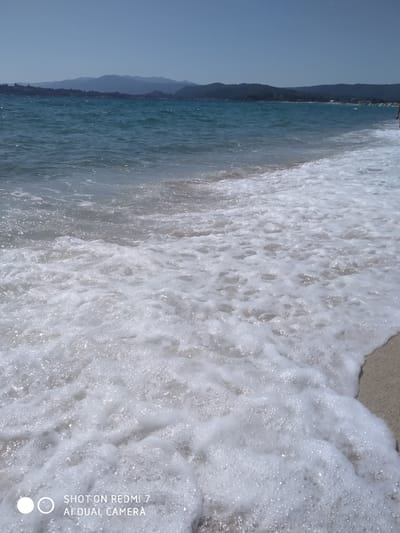 This image captures a serene beach scene in Sithonia, Greece. The foreground is dominated by frothy white waves gently rolling onto a sandy shore, with sunlight reflecting off the water's surface, creating a shimmering effect. In the mid-ground, the turquoise waters of the sea extend towards a distant coastline lined with lush green hills and mountains under a clear blue sky. The horizon is marked by the subtle silhouettes of the mountains, suggesting a peaceful, natural landscape. The overall impression is one of tranquility and natural beauty, typical of the Sithonia peninsula.