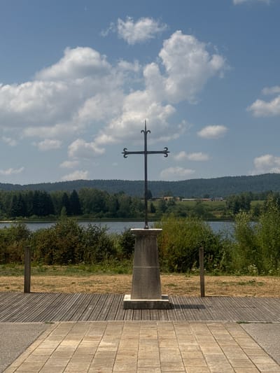 In Grande-Rivière Château, France, a dark metal cross stands prominently on a stone pedestal. The cross features decorative scrollwork at the ends of its horizontal bar. It is positioned in the foreground, with a wooden boardwalk extending to its left and right. Beyond the boardwalk, dry grass and dense green bushes are visible. In the mid-ground, a body of water, likely a lake or river, stretches horizontally, bordered by a line of diverse green trees. The background consists of rolling hills covered in a dense forest, with a few buildings nestled on the slopes. The sky above is a clear blue with scattered white clouds.