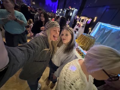 An upside-down photo shows a crowded indoor space. Two women stand in the center, one sticking her tongue out. To the left, a table is decorated with a gold cupcake wrapper. Behind it, a display of photos and lights hangs against a dark blue backdrop. People are congregated in the background, some wearing winter clothing. The lighting is dim, with blue and white lights accentuating the room's decor.