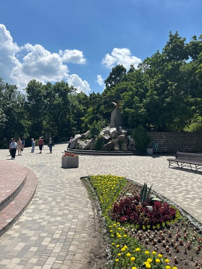 The image depicts a park in the city of Zheleznovodsk, Russia, on a sunny day with a blue sky and scattered white clouds. The foreground features a paved walkway with a checkered pattern of gray and beige tiles, bordered by a flower bed containing yellow marigolds and dark red plants. To the left, a curved red brick structure, possibly stairs or a low wall, is visible.
Further back, several people are walking on the paved area. Among them are individuals wearing casual clothing, some walking in groups and others individually. A large grey planter box with red flowers is positioned in the middle ground. Beyond the walkway, a rock formation serves as a base for a golden statue of an eagle with its wings spread. Behind the rock formation, a dense arrangement of green trees forms a backdrop. To the right, a stone wall runs along the background, with a small blue birdhouse-like structure and a wooden park bench visible near it. The overall scene suggests a public park or garden area designed for recreation and appreciation of nature.